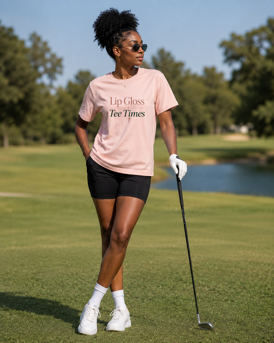 Woman wearing Lip Gloss & Tee Times women's golf tee with black shorts and golf club on golf course
