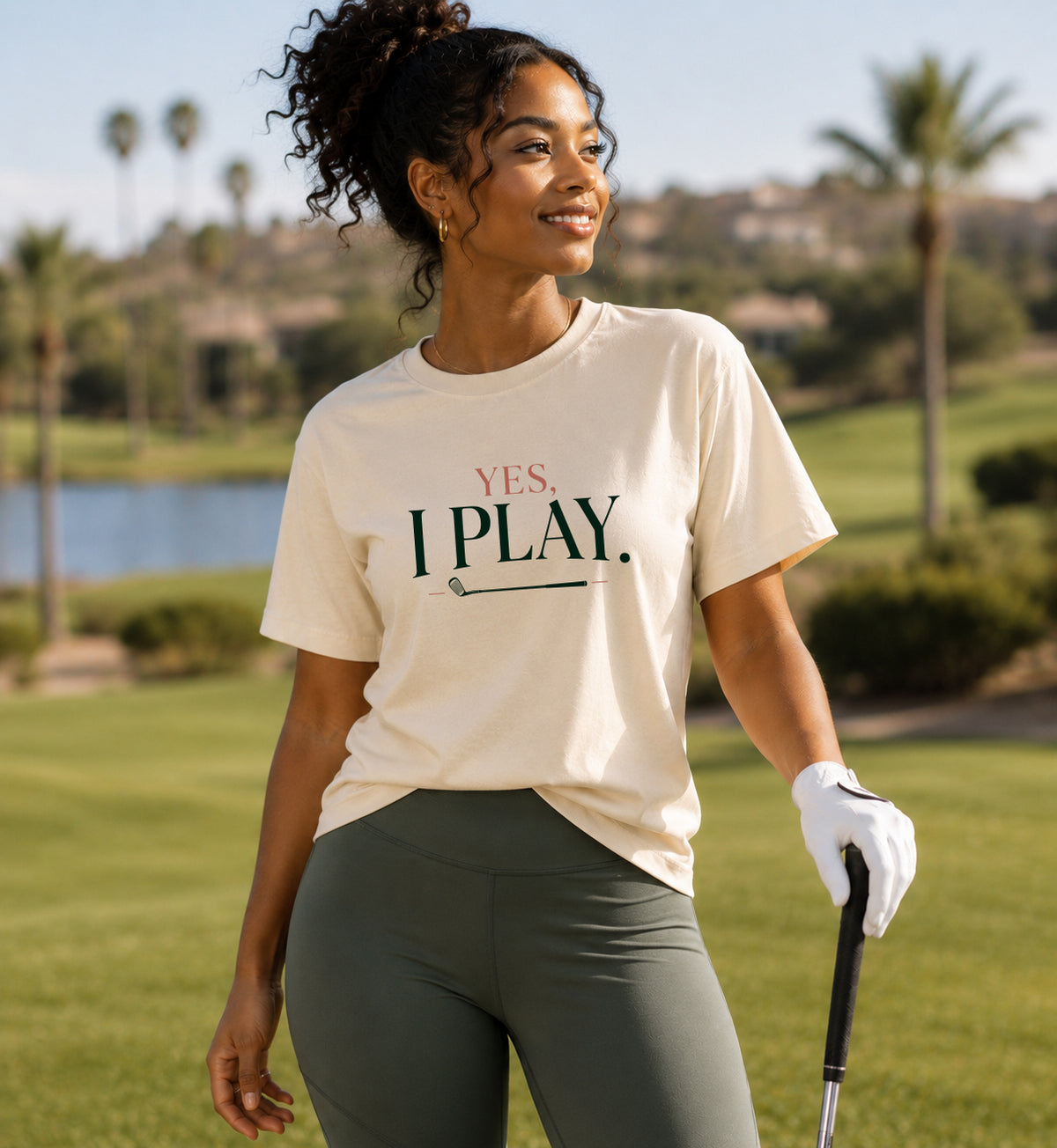 Woman wearing Yes. I Play. golf tee with gray pants and golf club on course